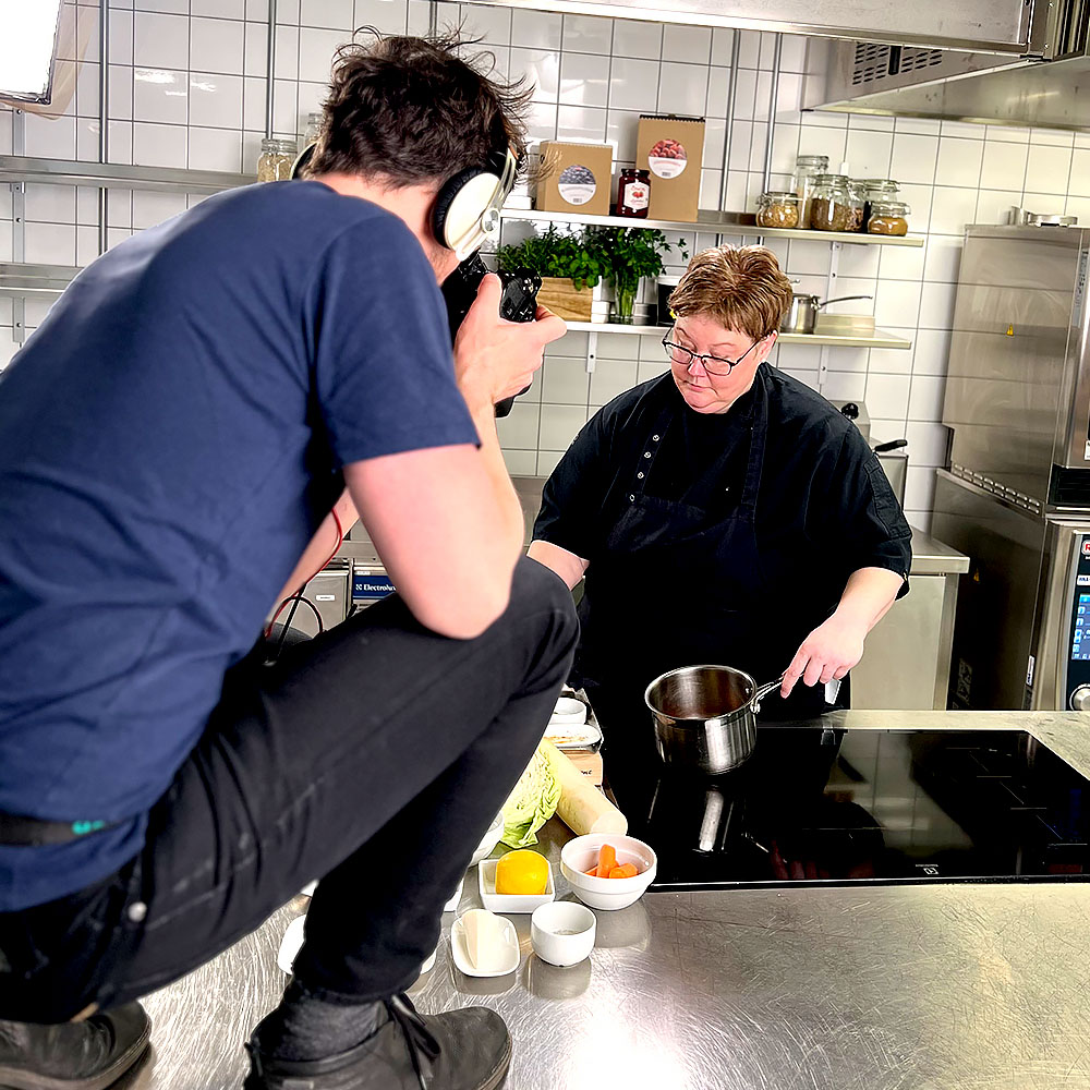 Filmare sitter på huk uppe på köksbänken med en kamera och filmar kocken som står vid spisen och håller i en kastrull.